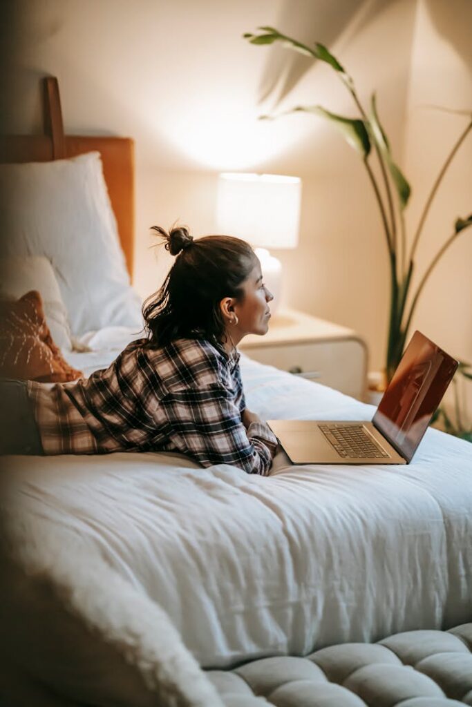 Side view of dreamy woman in checkered shirt lying on comfortable bed with laptop while working remotely in cozy bedroom