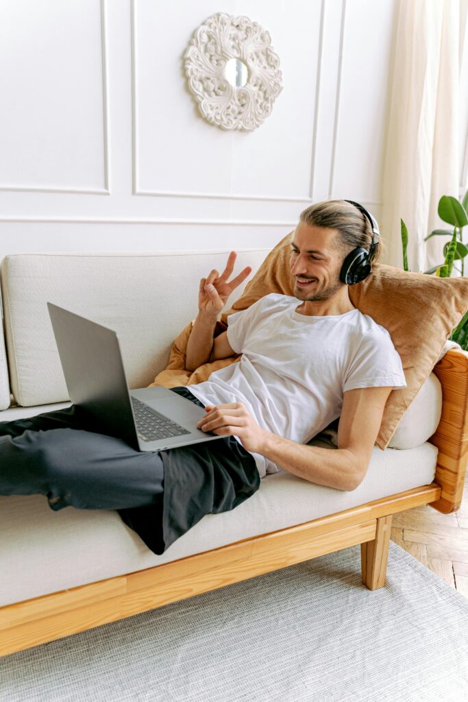 pexels photo 6598978 6598978 A man in a white shirt enjoys a relaxed moment with headphones and a laptop on a cozy sofa.